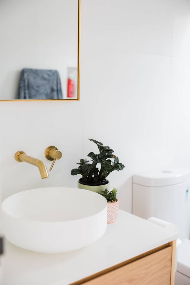 Bright contemporary bathroom vanity with a round white vessel basin, wall-mounted brushed brass tapware, white tiled walls, and a timber-fronted vanity. Potted plants add greenery, while a large framed mirror and toilet are visible in the background.