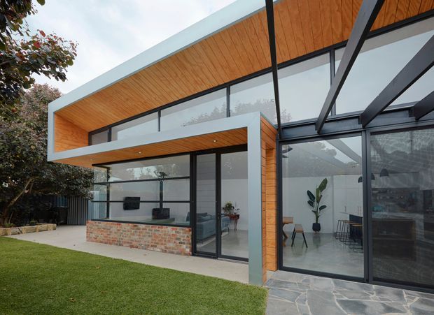 Exterior view of a contemporary single-storey home with strong geometric roof overhangs, extensive black-framed floor-to-ceiling glazing, warm timber soffits and wall cladding, and a low recycled-brick feature wall. The house opens onto a paved terrace and neatly landscaped lawn, with a bright minimalist interior visible through the glazing.