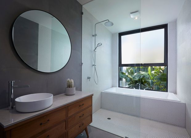 Contemporary bathroom featuring a freestanding round vessel basin on a timber vanity, large circular mirror, walk-in shower with overhead and handheld fittings, and a built-in tiled bath beneath a black-framed window. Small-format white textured tiles cover the shower and bath areas, creating a clean, bright interior with dark grey wall contrast and warm timber joinery.