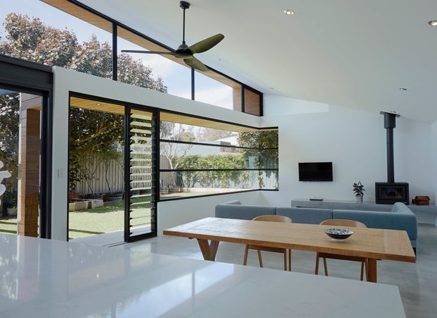 Bright, open-plan contemporary living and dining area viewed from a white kitchen benchtop. The space features extensive black-framed floor-to-ceiling glazing opening to a landscaped courtyard, a timber dining table, pale upholstered sofa, wall-mounted television, wood-burning fireplace with a tall black flue, and a large sculptural ceiling fan. White walls and polished pale flooring create an airy indoor-outdoor connection.