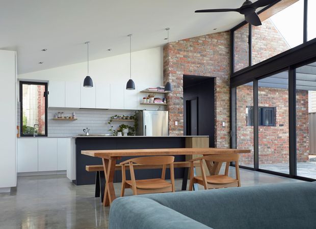 Open-plan contemporary kitchen and dining area with a visible living room foreground. The kitchen features white full-height cabinetry, a dark island, pale tiled splashback, stainless-steel appliances, and pendant lighting. A timber dining table and chairs sit beside the island, while a large black-framed glazed wall opens the space to an adjacent brick courtyard. Exposed red brickwork, polished concrete flooring, and a ceiling fan add material and visual interest.