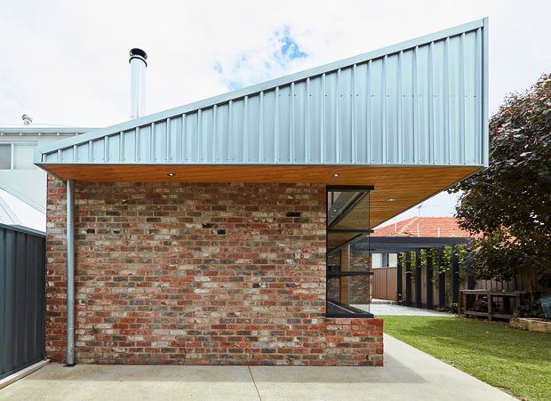 Exterior view of a compact contemporary brick-clad building with a dramatic asymmetrical pitched metal roof and broad timber-lined eave. The facade features exposed multicoloured brickwork, a tall narrow glazed opening with dark metal framing, and a simple concrete-paved approach. A small landscaped courtyard and adjoining residential structures are visible beyond.