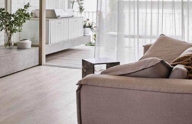 Bright, calm contemporary living room with a large taupe upholstered sofa in the foreground, sheer white curtains, pale timber-look flooring, and a minimalist white built-in cabinet visible through an open glazed door. Indoor plants and a small dark stone coffee table add natural contrast, while abundant filtered daylight creates an airy atmosphere.