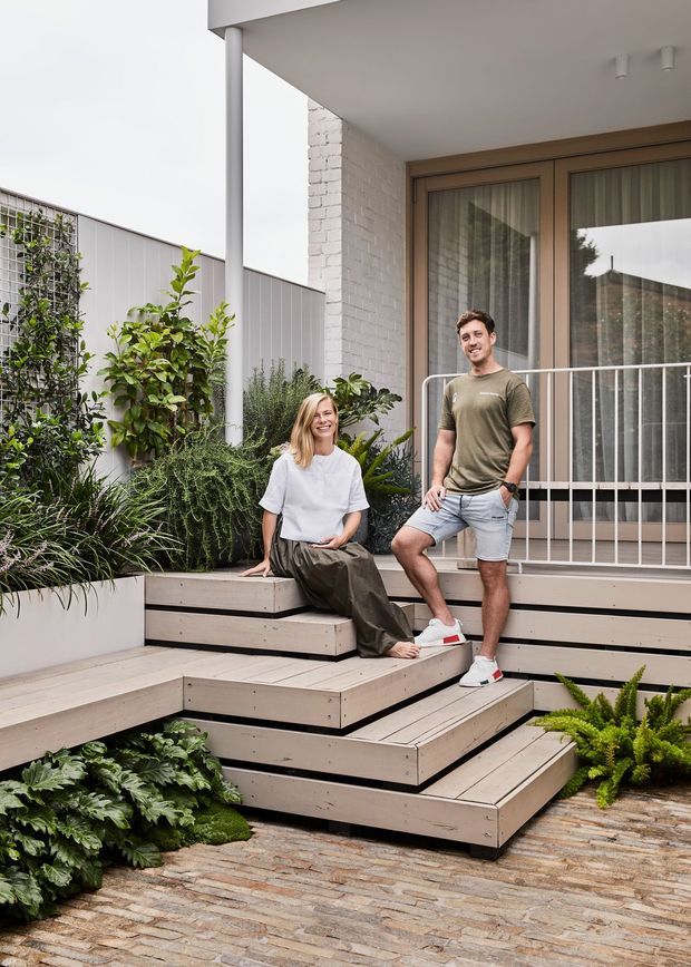 A contemporary residential entry terrace with broad tiered timber decking and steps leading to a raised front porch. The light-toned deck is integrated with built-in planter boxes filled with lush greenery, while a white-painted brick wall, timber-framed glazed doors, and a slim white metal balustrade define the facade. Two people are seated and standing on the steps. The foreground is paved in textured stone or brick, creating a landscaped, layered outdoor approach.
