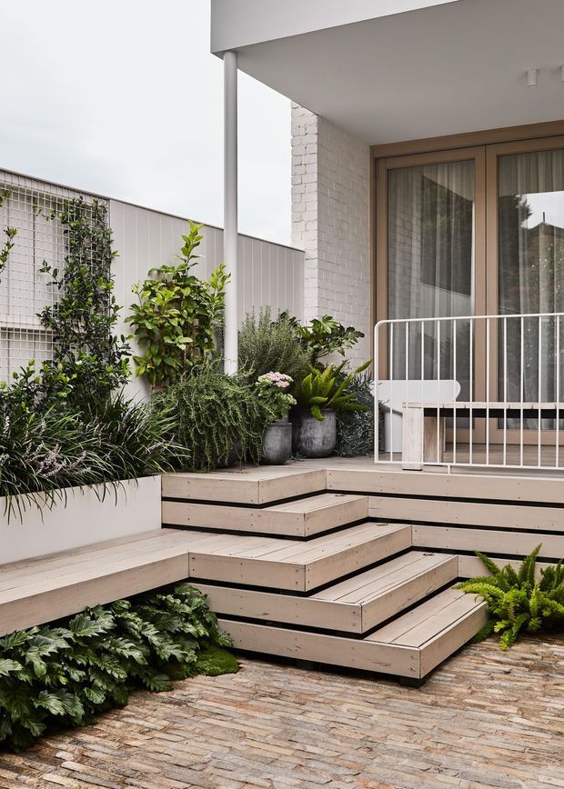 A landscaped residential courtyard or terrace entrance with tiered light timber decking and steps, integrated planter boxes, lush greenery, and a white painted brick wall. A covered porch with slim white posts, pale timber-framed glazed doors, and a simple white metal balustrade occupies the right side. The lower ground is paved with small rectangular stone or brick pavers, creating a layered contemporary outdoor space.