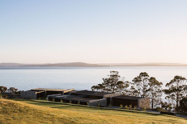 A low-profile contemporary home is nestled into a gently sloping grassy site beside a broad body of water. The building is composed of dark, rectilinear volumes with extensive flat roofs, dark window and door openings, and sections of rough grey stone cladding. Native-looking trees partially screen the house, while distant low hills sit across the water under a pale sky.