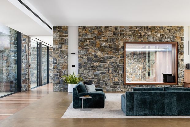 Contemporary living room with a dramatic exposed rough-stone feature wall, large timber-framed mirror, dark teal velvet seating, pale area rug, warm timber flooring, and expansive black-framed glazing along one side. The space is bright and refined, combining natural stone with minimalist modern furnishings.