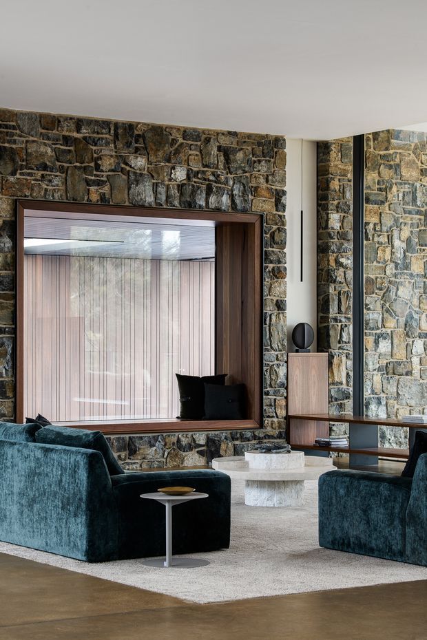 A refined living room with a dramatic exposed irregular-stone feature wall, large timber-framed interior opening, and warm vertical timber panelling visible beyond. Deep teal upholstered lounge chairs surround a low round pale stone coffee table, with a slim side table, low timber shelving, sculptural decor, and a neutral textured rug over a polished concrete or stone floor. The space has a low, clean ceiling and carefully integrated contemporary joinery.