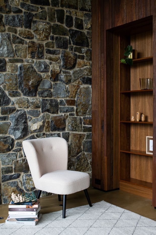 A moody, refined interior corner featuring a full-height rough natural stone wall beside dark timber wall panelling and built-in open shelving. A light upholstered lounge chair sits on a pale geometric rug over a dark floor, with stacked books and a chess set nearby. The timber shelving displays small decorative objects and a plant, creating a warm library or living-room reading nook.