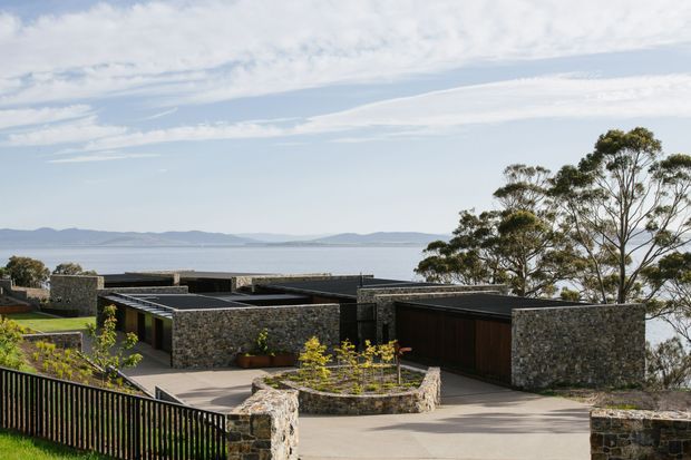A low-profile modern residence stepping down a sloped site toward a broad body of water, with dark flat roof forms, extensive rough stone walls, timber-toned garage doors, paved courtyards, and landscaped garden beds. Mature trees frame the view, while distant hills and calm water create a coastal or waterfront setting.