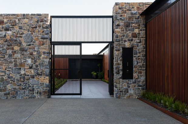 Contemporary residential entry courtyard viewed from the driveway, framed by robust irregular stone masonry walls and a tall black metal gate with fine vertical bars. A dark timber-look vertical slatted screen forms the right-hand boundary, while pale large-format paving leads through the entry toward a sheltered internal courtyard with planting. The composition emphasizes strong geometric lines, contrasting natural stone, dark metal, and warm timber tones.