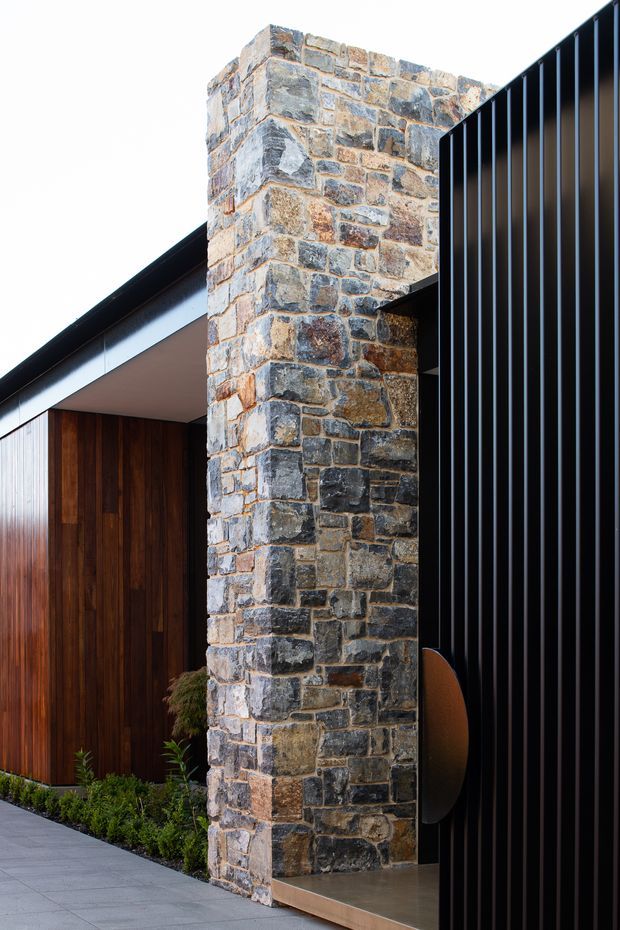 Close-up exterior architectural view showing a tall, rough-faced natural stone masonry pier beside a dark vertically ribbed metal facade. A warm timber-clad entry wall and roof edge are visible behind, with a paved approach and narrow planted strip at the base. The composition emphasizes contrasting stone, timber, and black metal materials in a refined contemporary residence.