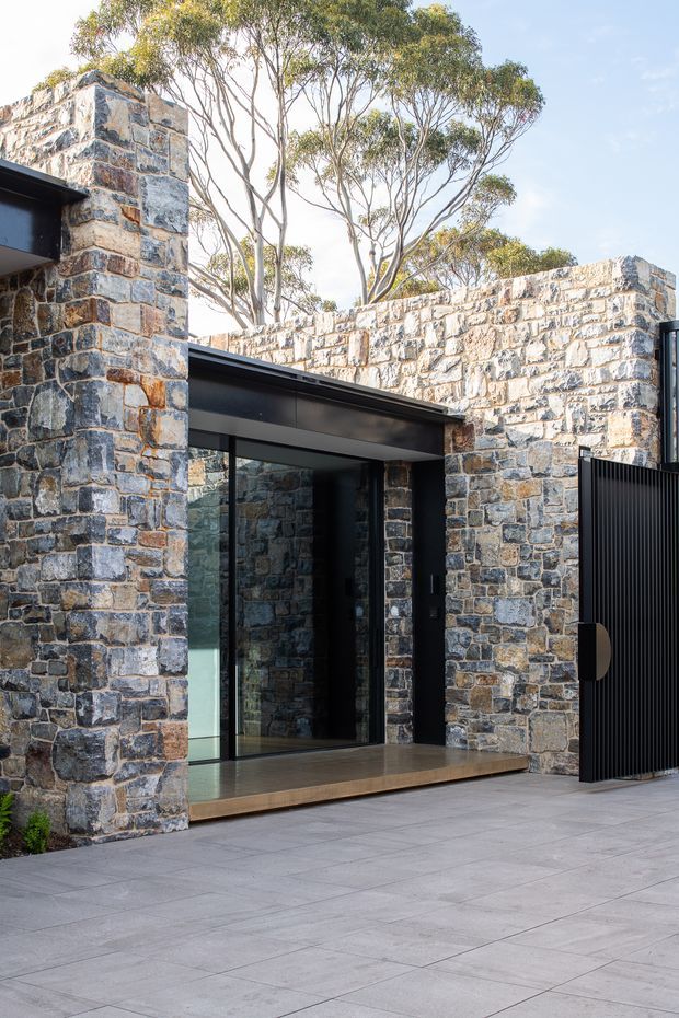 Exterior view of a contemporary residence centred on a recessed glazed entry beneath a dark flat roof. The facade and boundary walls are constructed from irregular multicoloured natural stone masonry, contrasted with black metal-framed glazing, a dark ribbed metal gate, and large grey-paved terrace slabs. Native eucalyptus trees rise behind the low stone walls.