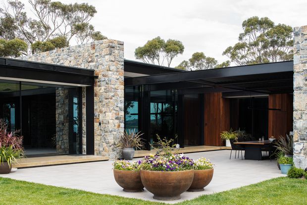 A contemporary single-storey courtyard-facing exterior with a low flat roof, dark metal-framed glazing, warm timber-clad recessed entry areas, and substantial natural stone masonry walls. A paved forecourt with bowl planters and garden planting leads into the building, with mature trees visible beyond.