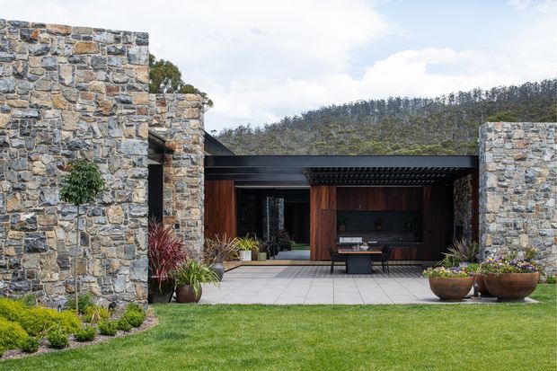 A low, contemporary residential courtyard viewed from the lawn, framed by substantial rough-faced natural stone walls. Dark metal flat-roofed or deeply recessed roof elements, warm timber-clad entry and outdoor living joinery, pale tiled paving, and landscaped planting create a strong connection between the building and garden. A wooded hillside is visible beyond the property.
