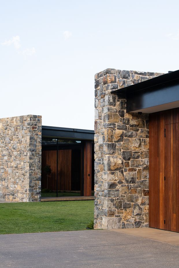 Exterior architectural photograph of a contemporary low-profile residence arranged around a grassy courtyard. The composition features robust irregular natural-stone walls, dark steel roof and beam elements, extensive dark-framed glazing, and tall warm timber entry doors. A paved forecourt occupies the foreground, with clean horizontal rooflines and strong geometric massing against an open sky.