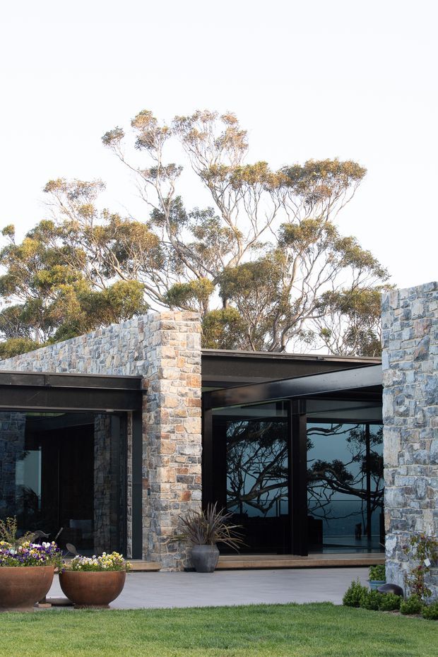 Exterior view of a contemporary, low-profile residence set among mature trees. The facade combines irregular natural stone walls with dark metal roof and structural framing, extensive floor-to-ceiling glazing, and a paved terrace bordered by large planters and manicured lawn. Reflections in the glazing show the surrounding trees and a distant blue landscape.