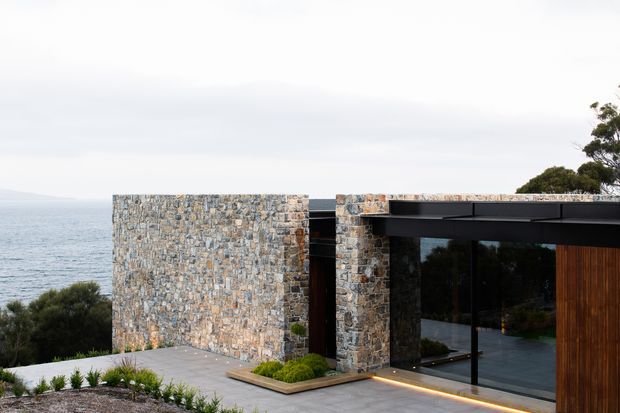 Contemporary coastal house exterior with low, flat-roofed forms clad in textured natural stone and dark metal framing. Full-height glazing opens onto a paved terrace, with subtle integrated lighting and landscaped planting; ocean and coastal vegetation are visible beyond.