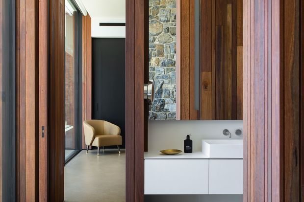 A refined contemporary bathroom vanity viewed through a richly timber-lined passage. The image features floor-to-ceiling dark timber wall and door panels, a minimalist white floating vanity with integrated basin, brushed metal wall-mounted tapware, and a small stone feature wall. An adjacent corridor with glazing and a mustard upholstered chair is visible beyond the timber opening.
