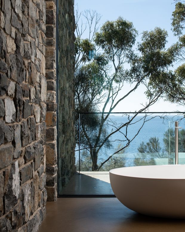 A contemporary bathroom with a freestanding white oval bathtub positioned beside full-height glazing overlooking blue water and coastal vegetation. A rugged exposed stone wall dominates the left side, contrasting with the smooth dark floor, frameless glass balustrade, and minimalist bathing fixture. The image has a refined, tranquil coastal atmosphere.