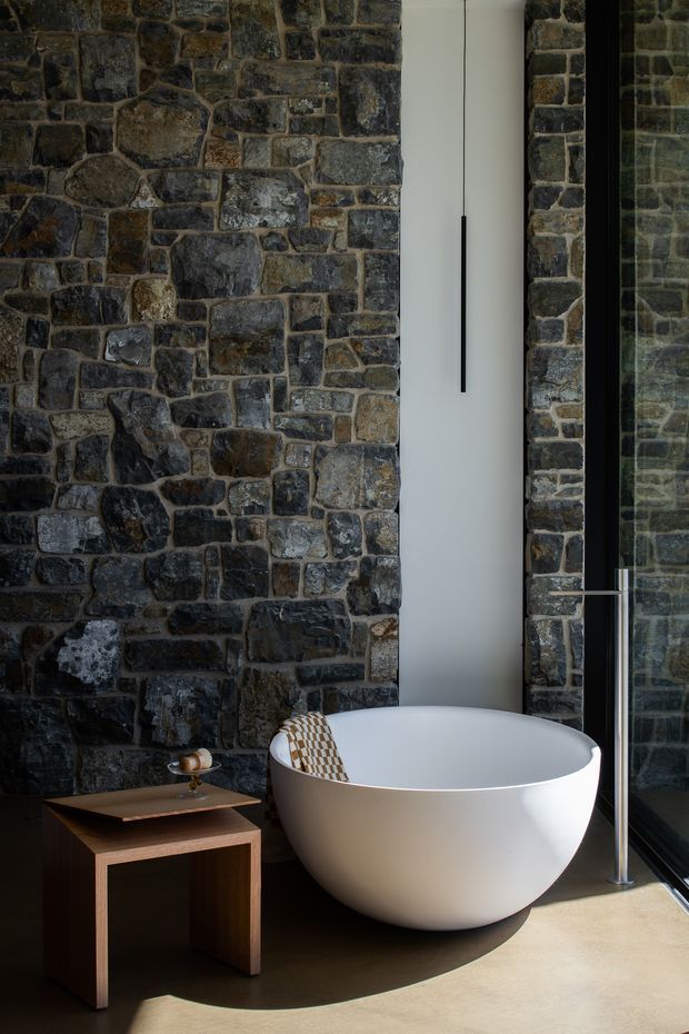 A dramatic, contemporary bathroom featuring a freestanding white oval bathtub positioned beside full-height dark-framed glazing. Rough, irregular dark stone masonry covers the walls, contrasting with a smooth pale floor and a minimalist black pendant light. A small timber side table with a copper-toned vessel sits beside the bath, creating a refined yet earthy atmosphere.