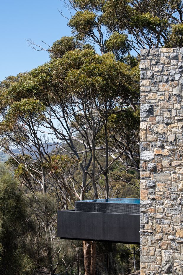Exterior architectural detail showing a dark cantilevered balcony or terrace with a circular spa/pool edge, attached to a substantial rough stone masonry wall. The structure is set among native eucalyptus trees on a wooded, sloping site, with distant hills visible through the vegetation under a clear blue sky.