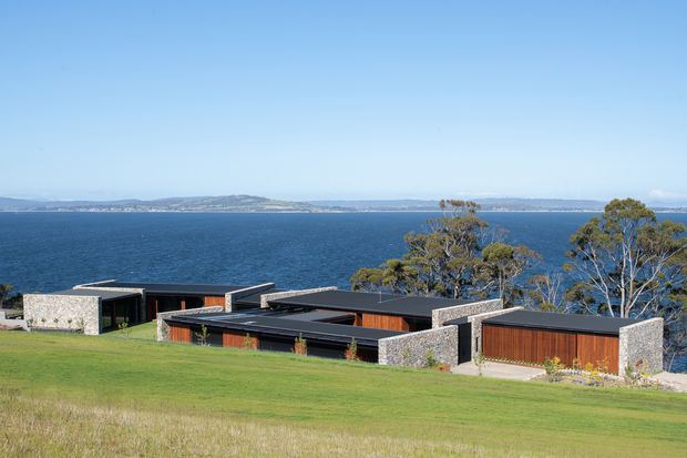 Low-profile contemporary waterfront residence set into a broad grassy site, with dark flat-roofed volumes, stone masonry walls, timber-clad sections, and expansive views across the water. Mature trees frame the house, while a distant shoreline is visible beyond.