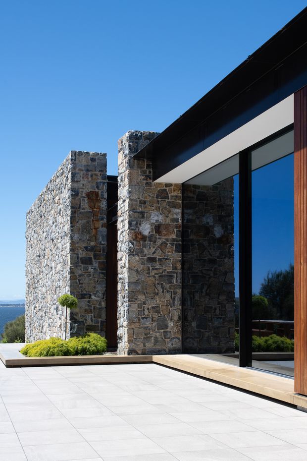 A contemporary residential exterior viewed from a broad paved terrace. The building combines rugged irregular natural-stone masonry walls and projecting stone piers with dark metal roof trim, large black-framed floor-to-ceiling glazing, and a vertical timber-clad section. Low sculpted planting borders the pale tiled terrace, with a distant blue landscape visible beyond.