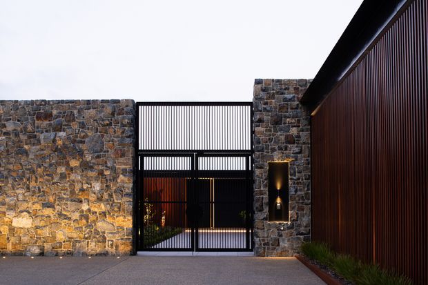 A contemporary residential entrance courtyard framed by rugged stacked-stone walls and a tall black metal slatted gate. Warm concealed lighting highlights the stonework and a narrow vertical wall feature, while a dark timber-toned vertical slat facade runs along the right side. The foreground is a paved driveway or entry court with narrow linear planting at the edge.