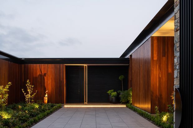 A dramatic contemporary residential entry courtyard viewed from outside, with a broad grey tiled walkway leading to a dark entrance door. The enclosed facade is clad in rich vertical timber, accented by dark metal framing and a prominent rough stone-clad vertical wall. Low planted garden beds and concealed ground lighting create a warm, refined arrival sequence beneath an overcast sky.
