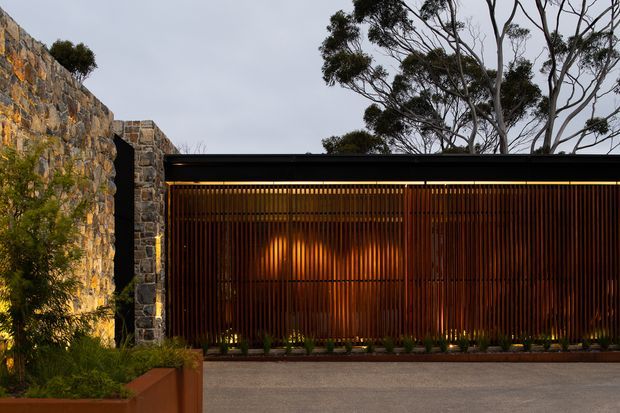 A contemporary residential facade or entry structure photographed at dusk, combining dark vertical timber battens with warm concealed lighting, a thin flat roofline, and rugged natural stone walls. Landscaping and mature trees frame the entrance, creating a refined connection between modern detailing and a natural site.