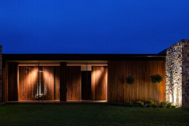 Night-time exterior view of a low, single-storey contemporary building with a dark flat roof, extensive warm timber wall cladding, recessed glazed openings, and a contrasting rough natural-stone wall at the right. Warm architectural lighting highlights the timber facade and entry glazing, with a dark lawn and deep blue sky in the foreground and background.