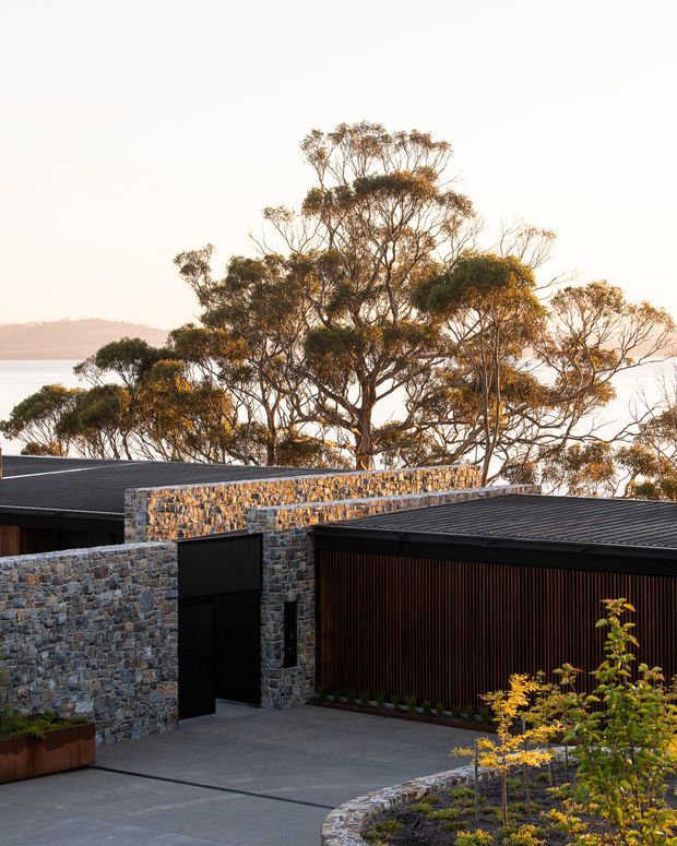 Exterior view of a contemporary low-profile residence with dark metal roofs, rugged stone masonry walls, a black pedestrian entry door, and a large timber-slatted garage or screening facade. A paved forecourt and curved planted garden bed sit in front, with mature eucalyptus trees and a hazy water or valley landscape beyond.