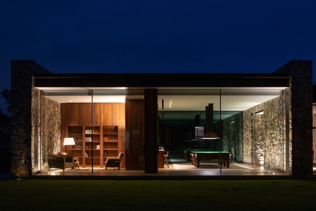 Night-time view of a sophisticated single-storey modern pavilion with extensive floor-to-ceiling glazing. Rough natural-stone walls and dark masonry end walls frame warm timber-lined interior spaces, including a reading or sitting area on the left and a billiards or games area on the right. Strong indoor-outdoor transparency and concealed linear lighting create a dramatic, refined atmosphere.