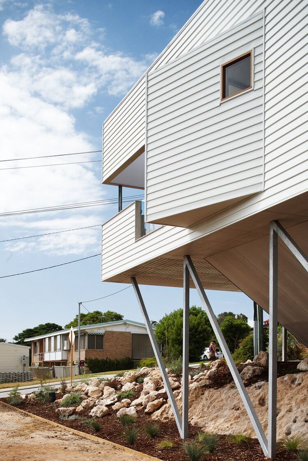 Exterior architectural photograph of a contemporary elevated house with white horizontal weatherboard cladding, projecting box-like volumes, and dramatic angled steel supports. The building sits on a rocky, landscaped sloping site, with a low-rise suburban context visible behind it under a bright sky.