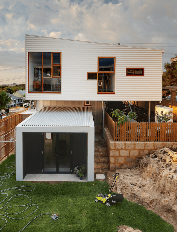 Exterior view of a compact contemporary two-storey dwelling on a sloping suburban site. The upper level is clad in pale horizontal weatherboards with large timber-framed windows, while a lower enclosed garage or studio volume has dark glazed doors and a corrugated metal roof. Timber fencing, exposed stone retaining walls, landscaped lawn, and ongoing excavation are visible in the foreground.