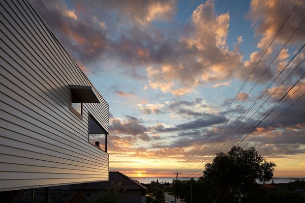 Exterior architectural photograph of a contemporary multi-level residential building at sunset. The facade is clad in horizontal pale weatherboards, with projecting dark-framed balconies or recessed openings. A suburban street, neighbouring rooftops, utility lines and silhouetted vegetation recede toward the glowing horizon.