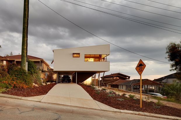 Street-level exterior view of a contemporary elevated house with a box-like upper storey clad in horizontal light-coloured weatherboards. The building is raised above a covered carport, with a broad concrete driveway, landscaped front garden, neighbouring homes, overhead utility wires, and a visible 20 km/h road sign. Warm interior light glows through the upper-level windows beneath a dramatic overcast sky.
