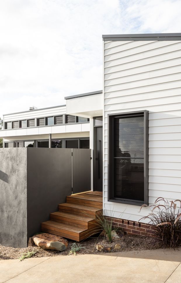 Exterior view of a contemporary residential dwelling with white horizontal weatherboard cladding, dark aluminium-framed glazing and trim, a flat roof, and a timber stair leading to a partially screened entry. A rendered boundary wall, dark metal gate, brick base course, and low ornamental planting complete the modern facade.