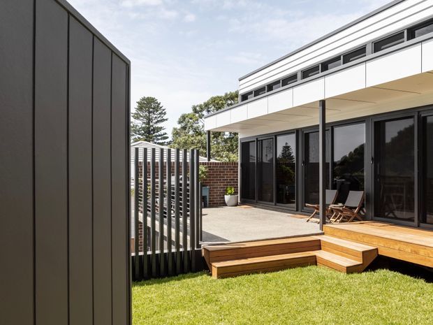 Contemporary residential exterior showing a low, flat-roofed building with white horizontal cladding, dark framed glazing, and a raised timber deck. The courtyard-like outdoor space includes stepped timber seating, a paved terrace, vertical metal screening, brickwork, and lawn.