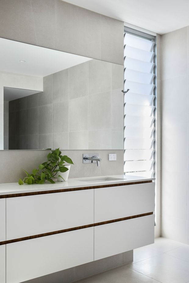 Bright contemporary bathroom vanity area featuring a floating white double-drawer cabinet with dark timber accent inlays, a white integrated basin, wall-mounted chrome tapware, large rectangular mirror, pale grey tiled wall surfaces, and a tall louvred window providing natural light. A small green plant sits on the vanity.