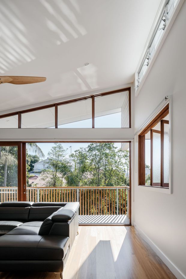 Bright, airy living room with a large black sectional sofa, polished timber flooring, white walls and a high sloping ceiling. Full-height timber-framed glazing and a glazed clerestory open onto a timber balcony, bringing in abundant sunlight and views of surrounding trees and neighbouring homes. A ceiling fan and strong daylight create a relaxed indoor-outdoor connection.