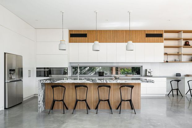 Bright contemporary kitchen with a large island and four bar stools. The space features white full-height cabinetry, a timber slatted overhead feature, a dramatic veined stone island benchtop and side panels, polished concrete flooring, stainless-steel appliances, open timber shelving, and pendant lighting. A dining or casual seating area is visible to the right.