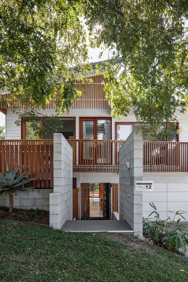 Front exterior view of a contemporary multi-level residence, partially screened by mature foliage. The facade combines light masonry block walls with warm vertical timber battens, timber balcony and entry gates, dark tiled paving, and large glazed openings with louvred window sections. A recessed pedestrian entrance sits between masonry walls, with the house number 12 visible beside it.
