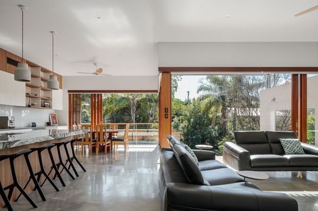 Open-plan contemporary living space combining a kitchen, dining area and lounge. The room features polished concrete flooring, extensive timber-framed glazing, built-in timber shelving and cabinetry, pendant lighting, a kitchen island with bar seating, a dining table, and a large black leather sectional sofa. Lush vegetation is visible outside through the glazed openings.