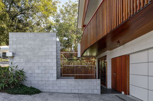 Contemporary residential exterior showing a recessed entry and garage beneath a cantilevered upper level. The facade combines warm vertical timber battens and soffit lining with pale grey concrete block walls, a slatted timber privacy screen, glazed doors, and a large white sectional garage door. Mature vegetation frames the setting.