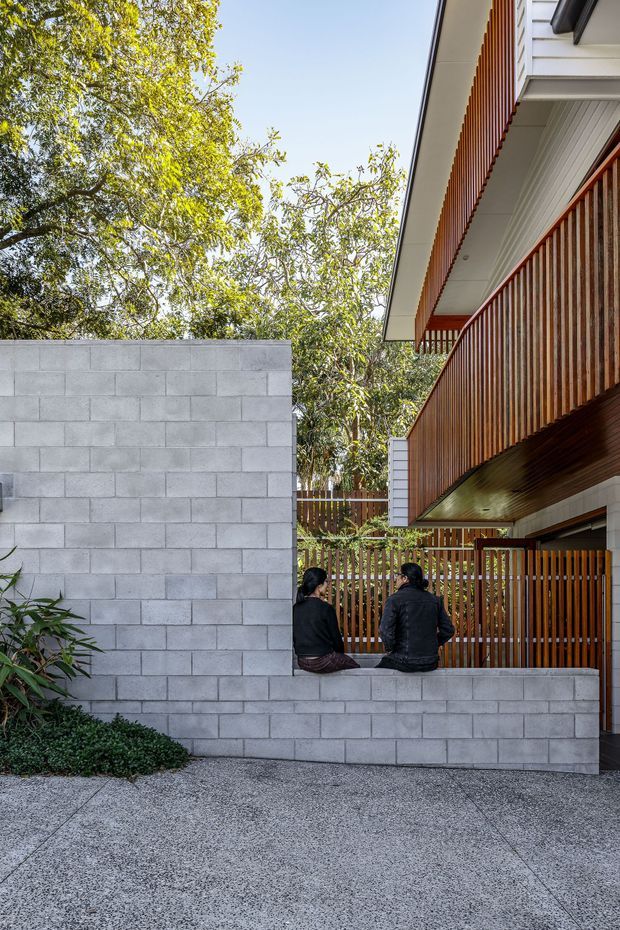 Exterior architectural view of a contemporary multi-level residence, showing a grey concrete block boundary wall, textured concrete paving, timber slatted balcony balustrades and screening, white eaves, and a sheltered entry or courtyard zone. Mature trees frame the building, while two people sit on the low boundary wall.
