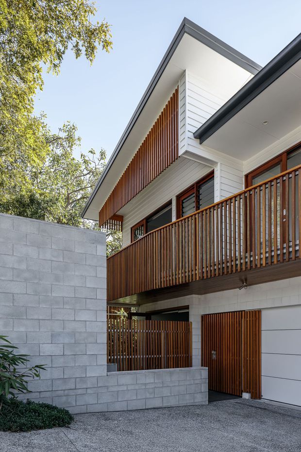 A contemporary multi-level residential facade viewed from the driveway, featuring white horizontal weatherboard cladding, a steep gabled roof, warm timber screening and vertical timber balcony balustrades, grey concrete block boundary walls, and a timber garage and entry door. Mature trees frame the property and the composition is bright and clean.