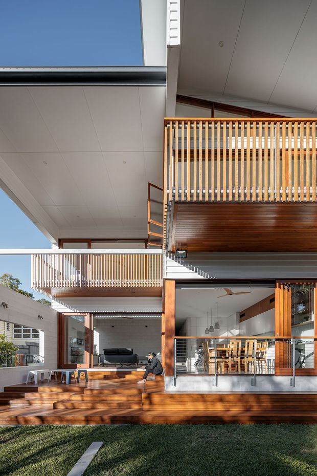 A bright, multi-level residential home viewed from the rear garden, with extensive timber decking, open-plan glazed living and dining areas, and stacked balconies enclosed by vertical timber balustrades. Large roof overhangs and a central open void create a strong indoor–outdoor connection. Warm timber framing and decking contrast with white soffits, pale walls, and dark metal details.