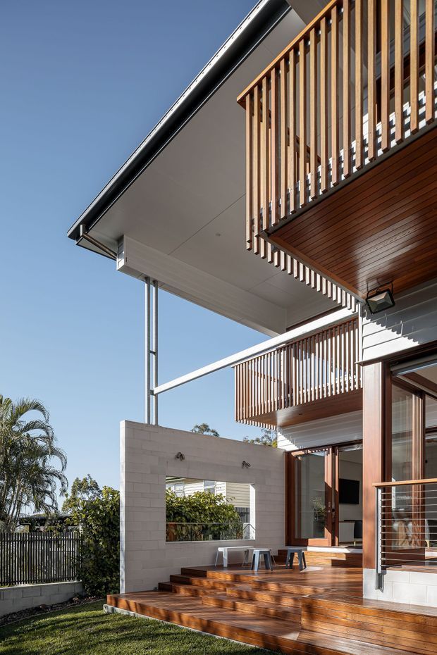 Exterior architectural photograph of a contemporary multi-level home with projecting upper floors, timber-slatted balcony balustrades, warm timber soffits and decking, white masonry walls, large glazed openings, and slender metal supports. The view focuses on the outdoor deck and facade under clear daylight, with landscaped planting visible around the property.